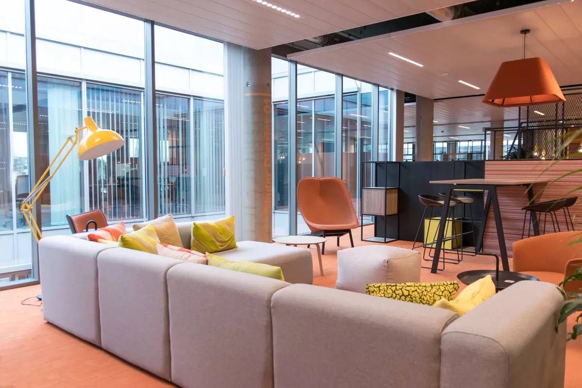 Un environnement de bureau ouvert avec un coin salon confortable, composé d'un canapé gris, de coussins colorés et de sièges 